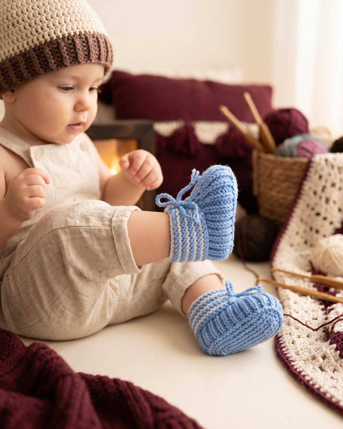 A baby wearing a cream linen romper, a beige and brown crocheted hat, and light blue crocheted booties sits on a white floor surrounded by knitting yarn and tools.