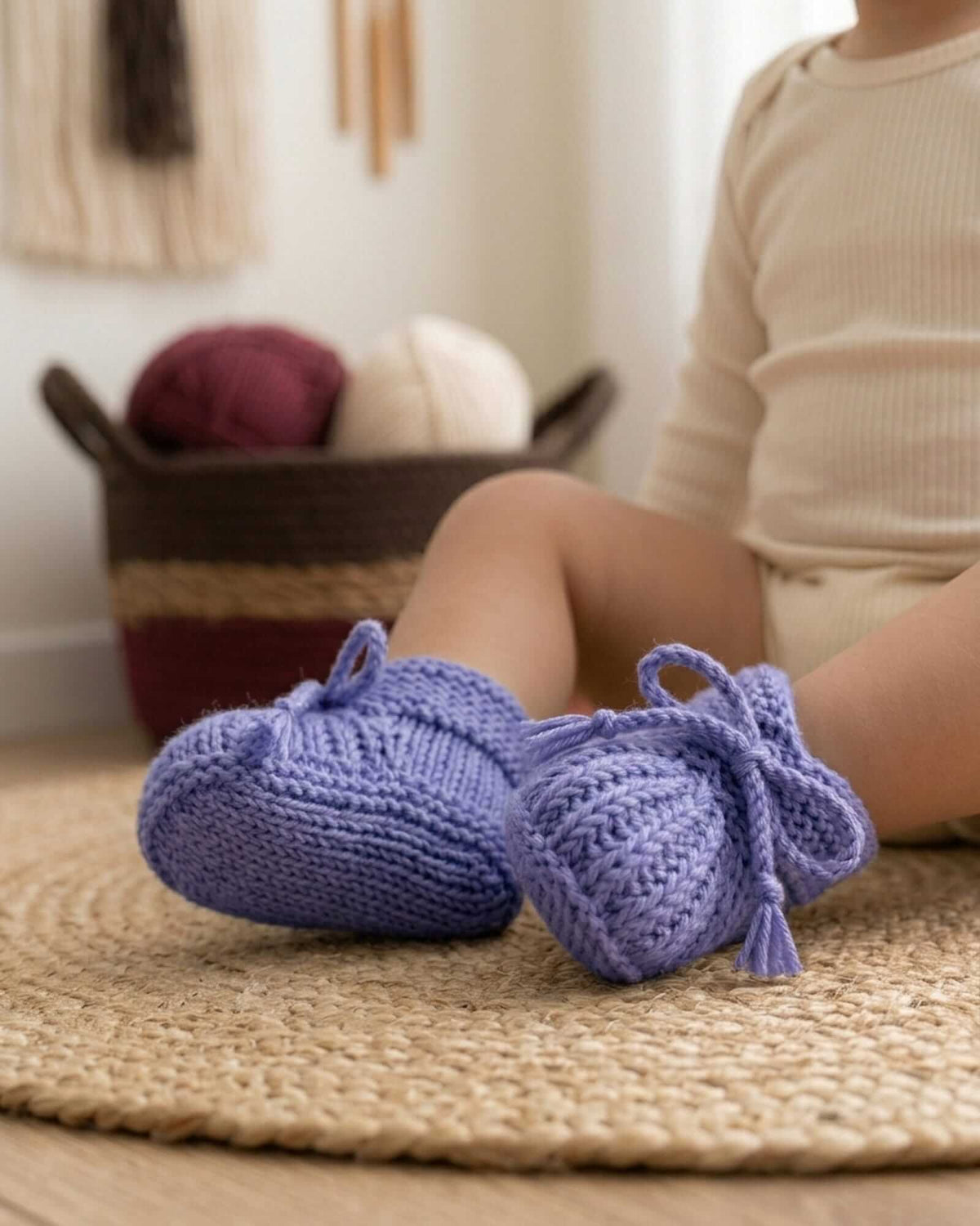 A pair of lavender hand-knit baby booties with tassel ties, shown on a baby sitting on a woven rug in a cozy room setting.