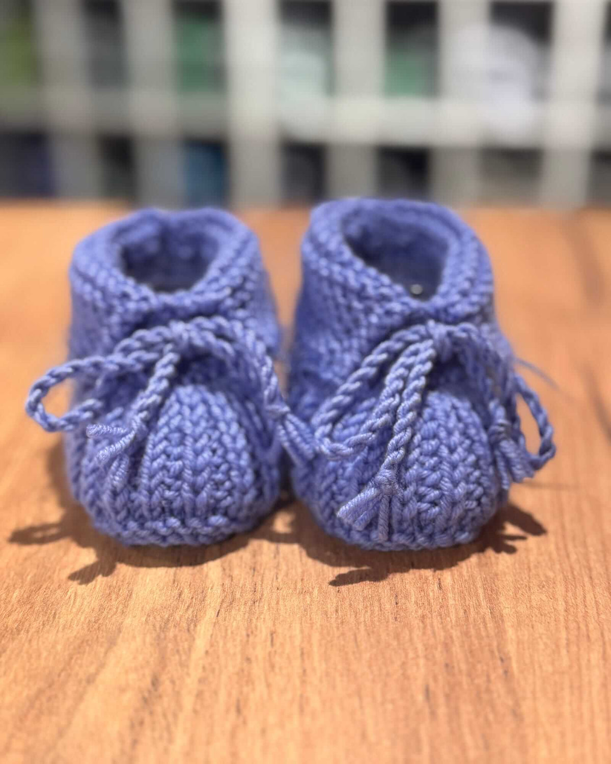 A pair of handmade, hand-knit baby booties in a dusty periwinkle blue, resting on a wooden tabletop with a blurred indoor background.