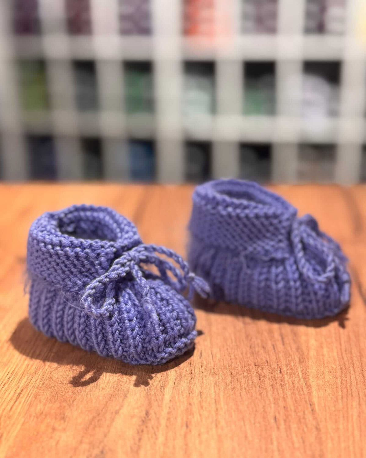 A pair of handmade, hand-knit baby booties in a dusty periwinkle blue, resting on a wooden tabletop with a blurred indoor background.