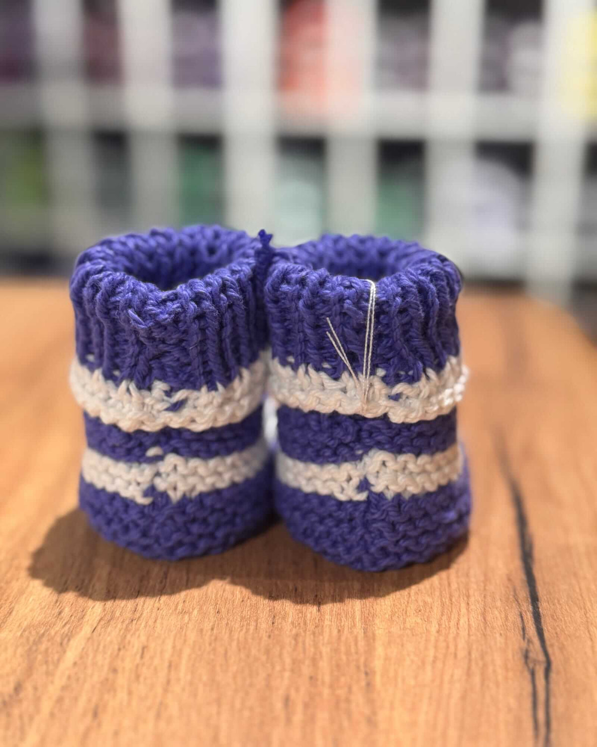 A pair of hand-knit baby booties in purple and cream stripes, resting on a wooden surface with a blurred background.