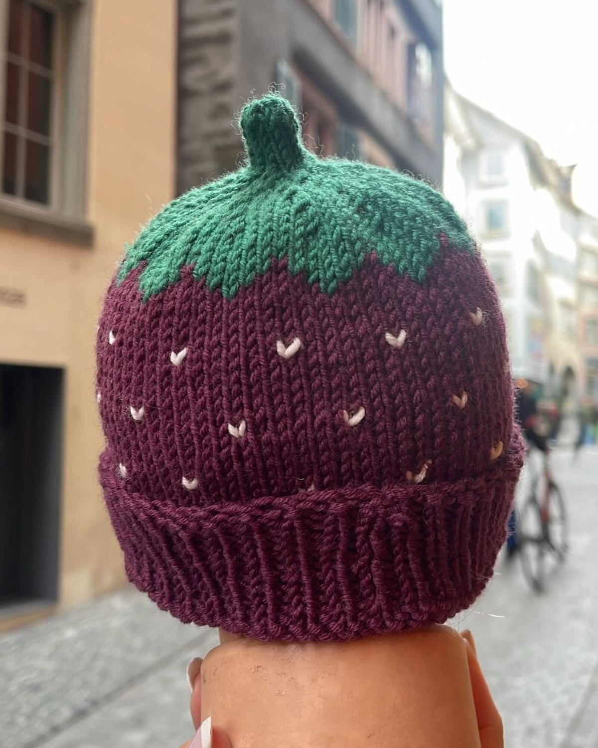 A hand-knit strawberry hat in burgundy and green yarn, shown from the back on a person's head in an outdoor street setting.