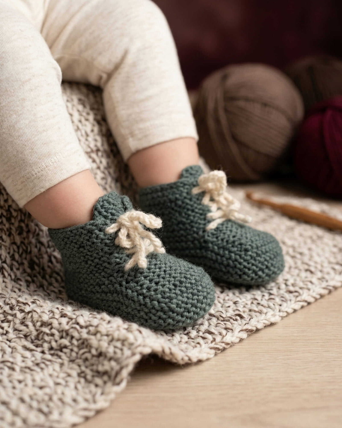A close-up of a baby's legs wearing handmade, dark green chunky-knit booties with cream-colored yarn bows, sitting on a beige knit blanket with yarn balls and a crochet hook in the background.