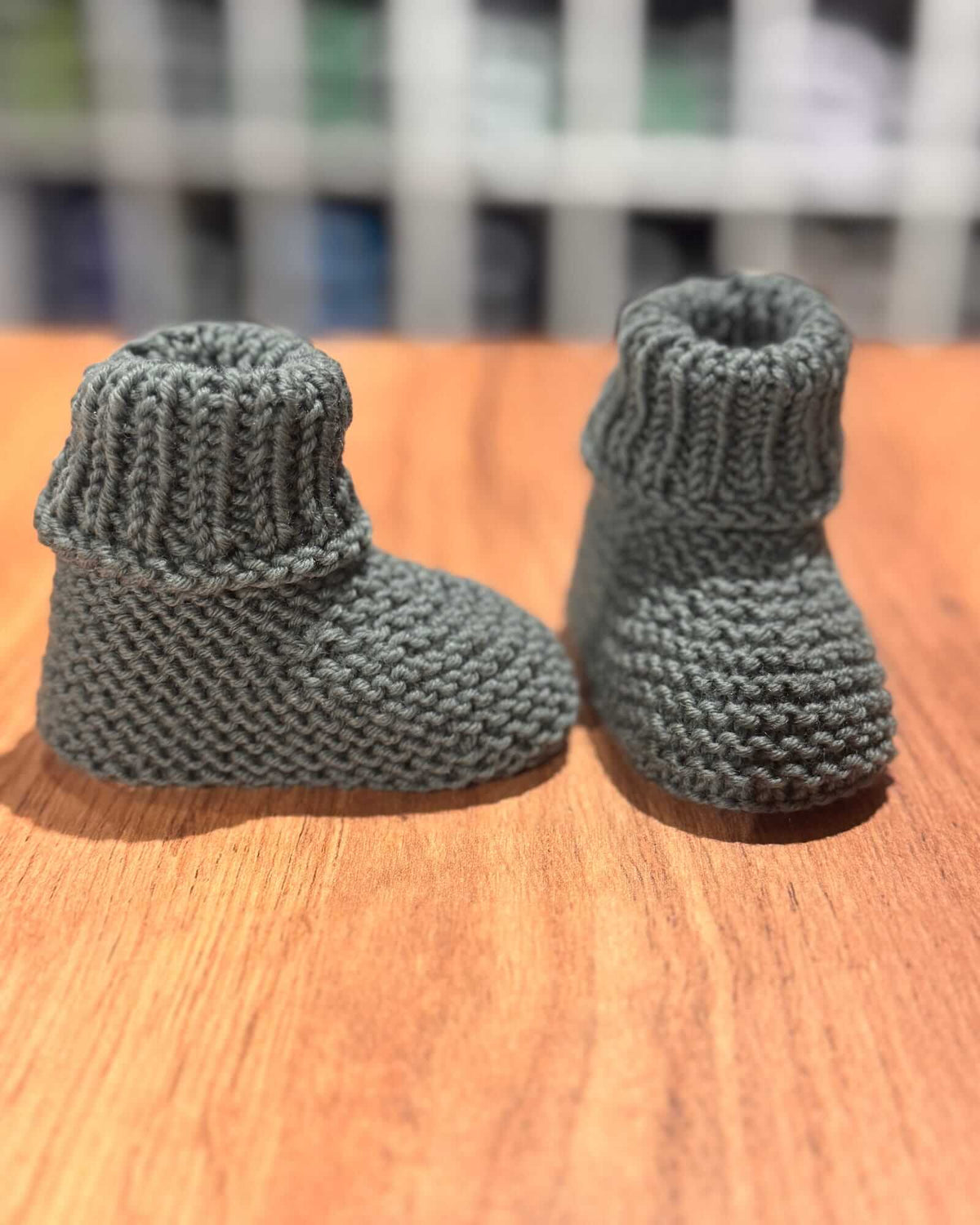 A pair of handmade, hand-knit baby booties in a muted gray-green color, displayed on a wooden surface with a blurred indoor background.