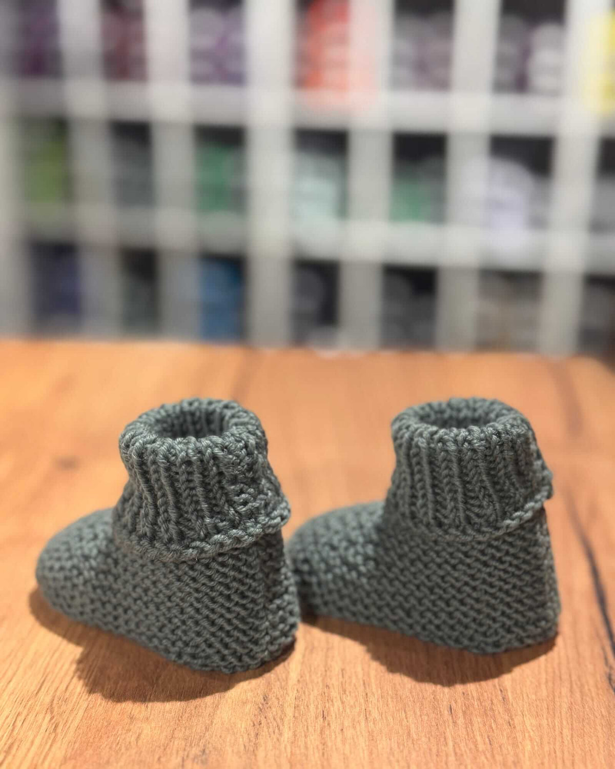 A pair of handmade, dark gray knitted baby booties resting on a wooden tabletop with a blurred background of shelves containing colorful yarn or thread.