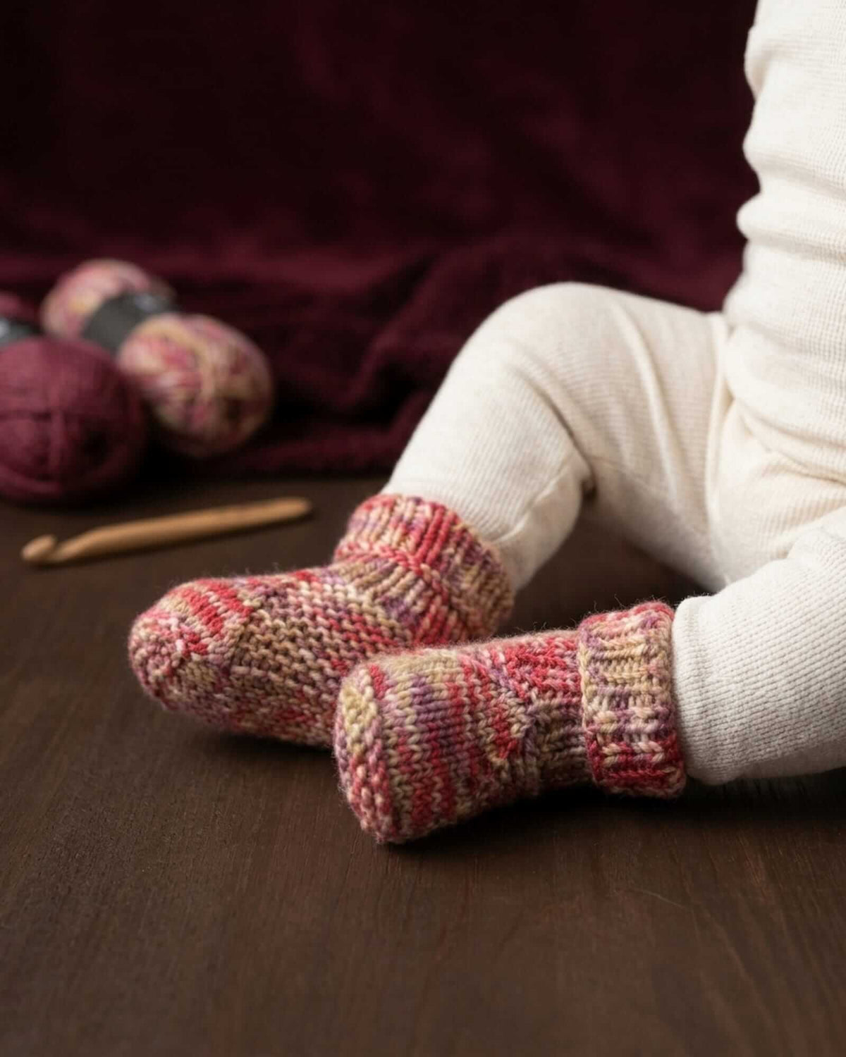 A close-up of a baby's legs wearing hand-knit baby booties in a red, pink, and beige pattern, sitting on a dark wooden floor with yarn balls and a crochet hook in the background.