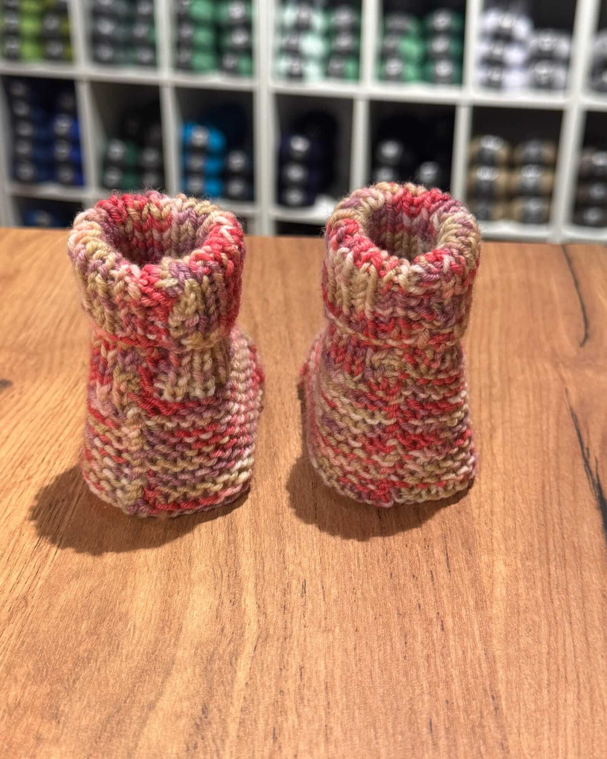 A pair of hand-knit baby booties in variegated yarn with red, pink, beige, and white colors, displayed on a wooden tabletop with a blurred store background.