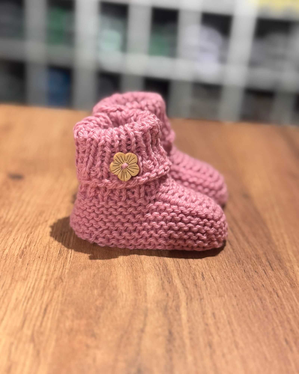 A pair of pink hand-knit baby booties with a small yellow flower accent on the front, resting on a wooden surface.