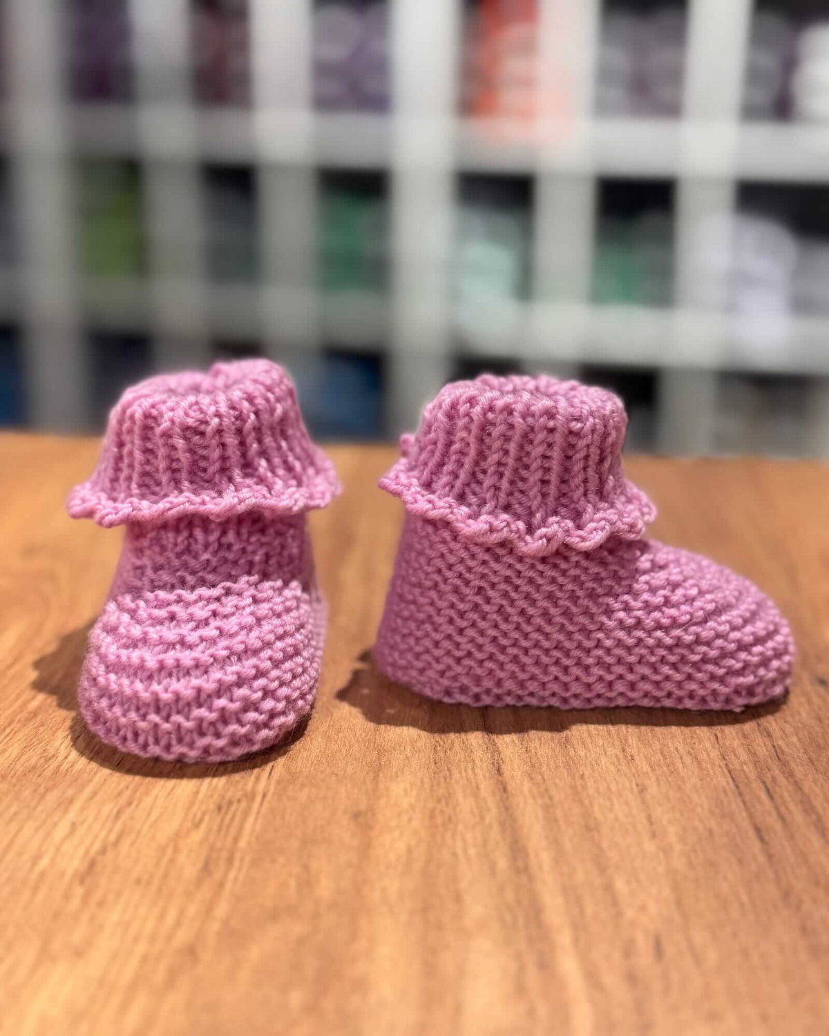 A pair of pink hand-knit baby booties with ruffled cuffs, displayed on a wooden surface with a blurred background.