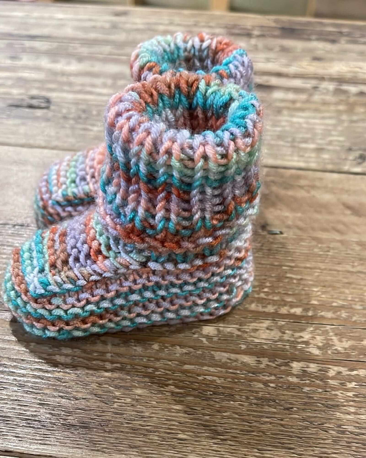 Hand-knit baby booties in a variegated pastel colorway of teal, peach, lavender, and white, displayed on a rustic wooden surface.