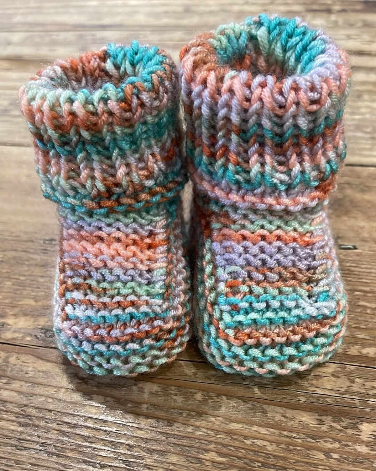 Hand-knit baby booties in a variegated yarn with stripes of teal, orange, pink, and lavender, displayed on a rustic wooden surface.