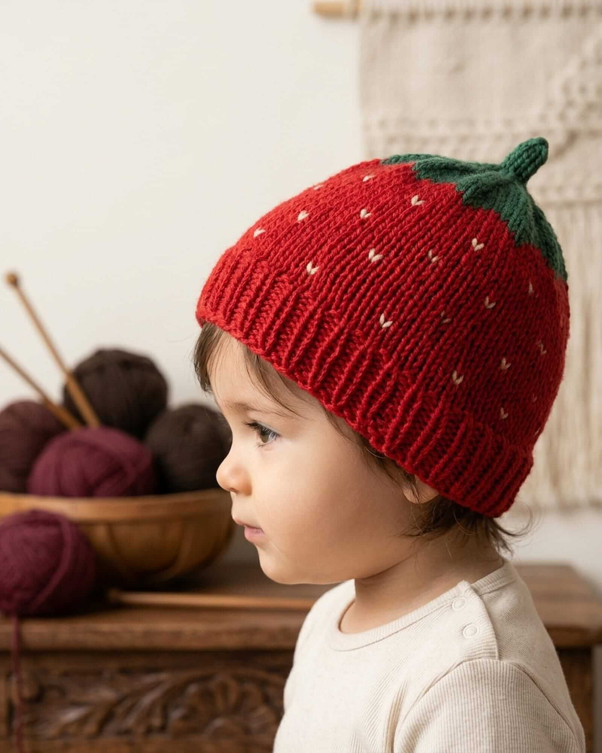 A child wearing a red strawberry-shaped knit beanie with green leaves and small white dotted details, photographed indoors with yarn balls and a wooden surface in the background.