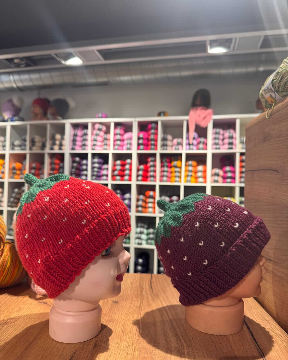 Two knitted strawberry-themed beanies displayed on mannequin heads on a wooden table, with a store shelf of colorful yarn in the background.