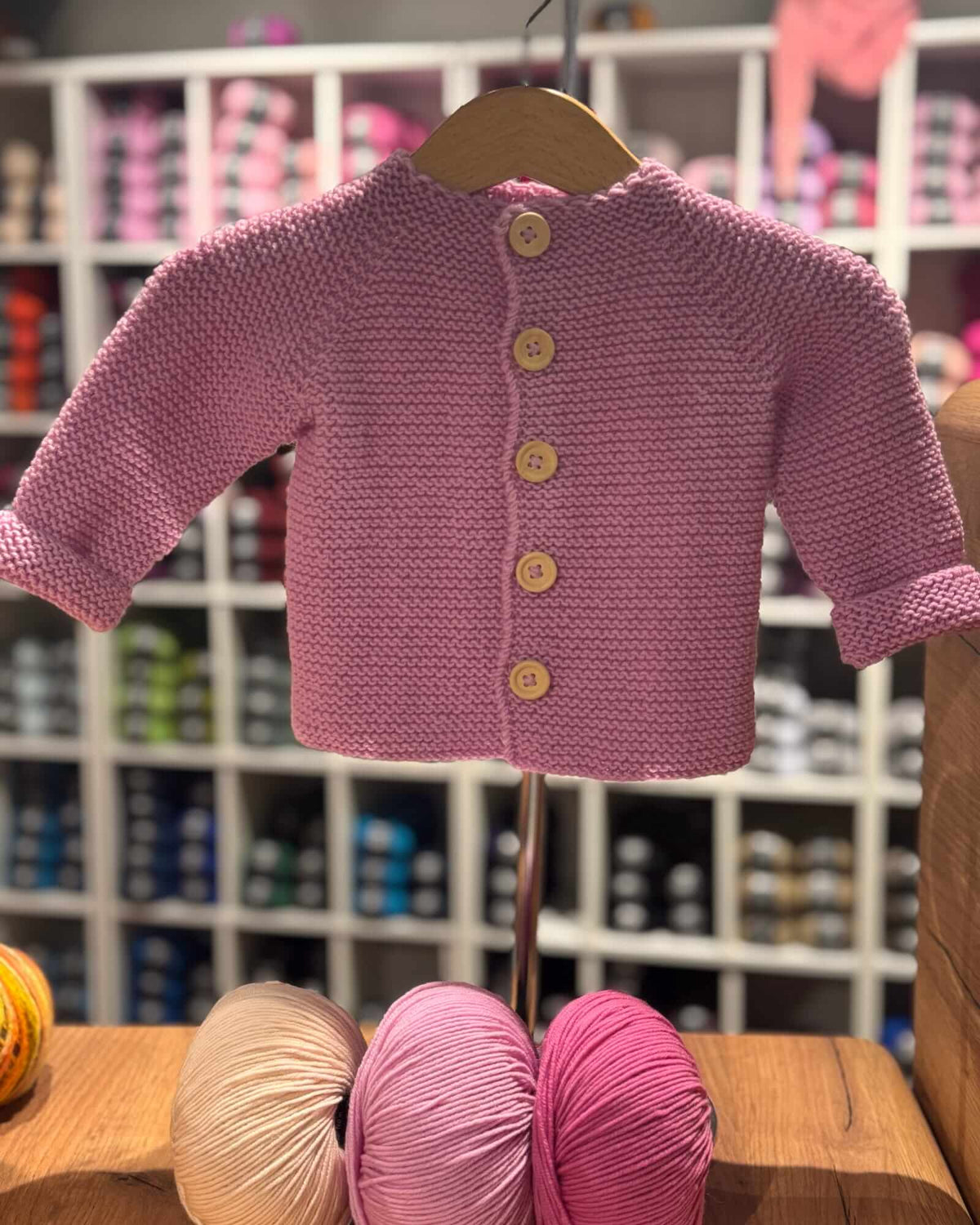 A pink knitted baby cardigan sweater with three-quarter sleeves and four wooden buttons, displayed on a wooden hanger on a table with yarn balls in the foreground and a store shelf in the background.