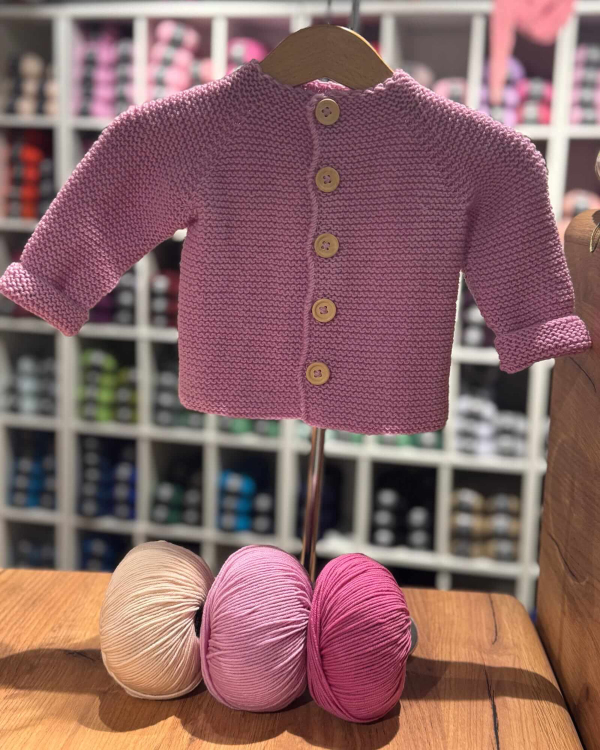 A small pink knitted cardigan or sweater displayed on a hanger stand, with three balls of yarn in beige, dusty pink, and magenta resting on a wooden table in front. The background shows a blurred store shelf filled with colorful yarn packages.