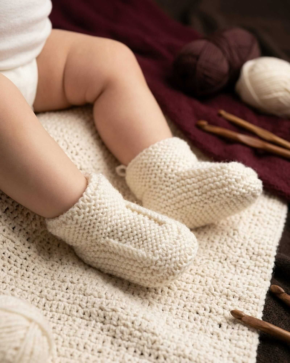 A close-up of a baby's legs wearing cream-colored, chunky-knit booties, sitting on a matching cream knit blanket with yarn and knitting needles in the background.