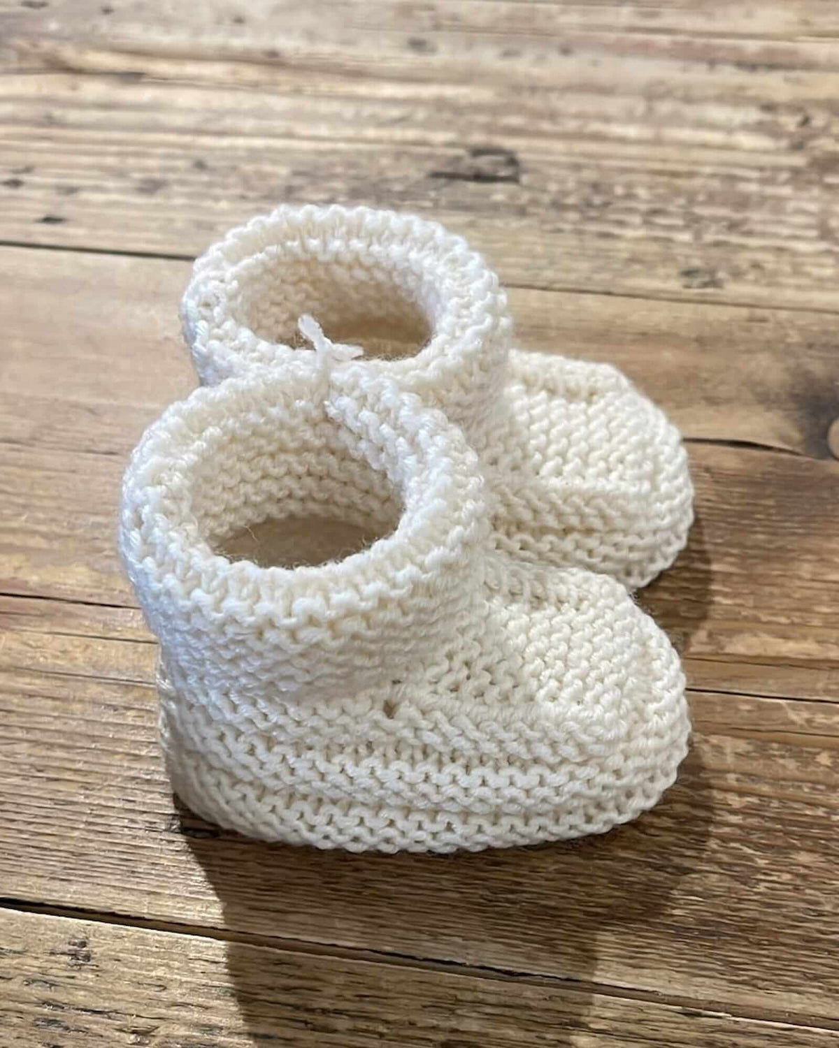 A pair of off-white hand-knit baby booties resting on a rustic wooden plank surface.