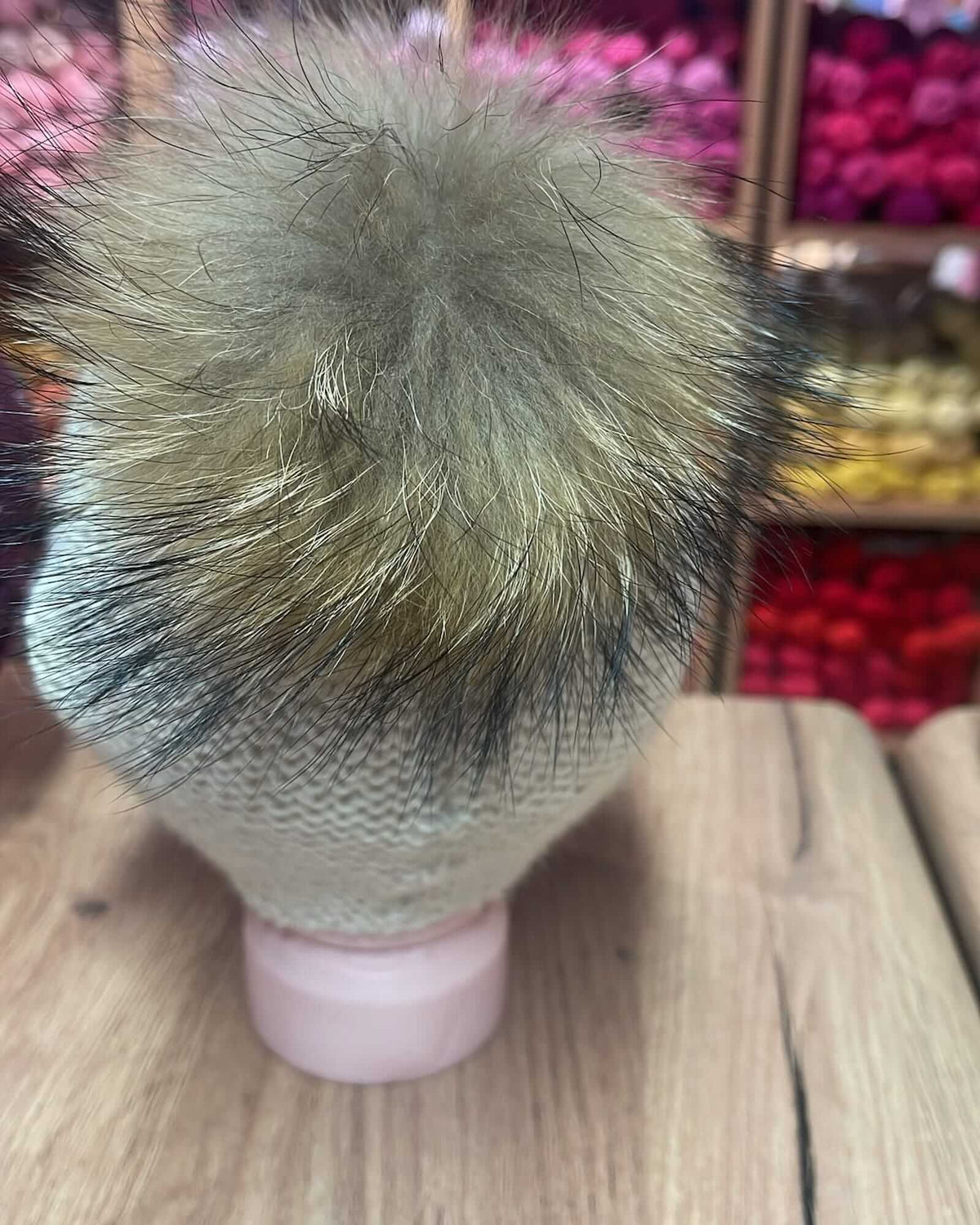 A close-up of a knit hat with a large fluffy pom-pom on a pink mannequin head, displayed on a wooden surface with a blurred shop background.
