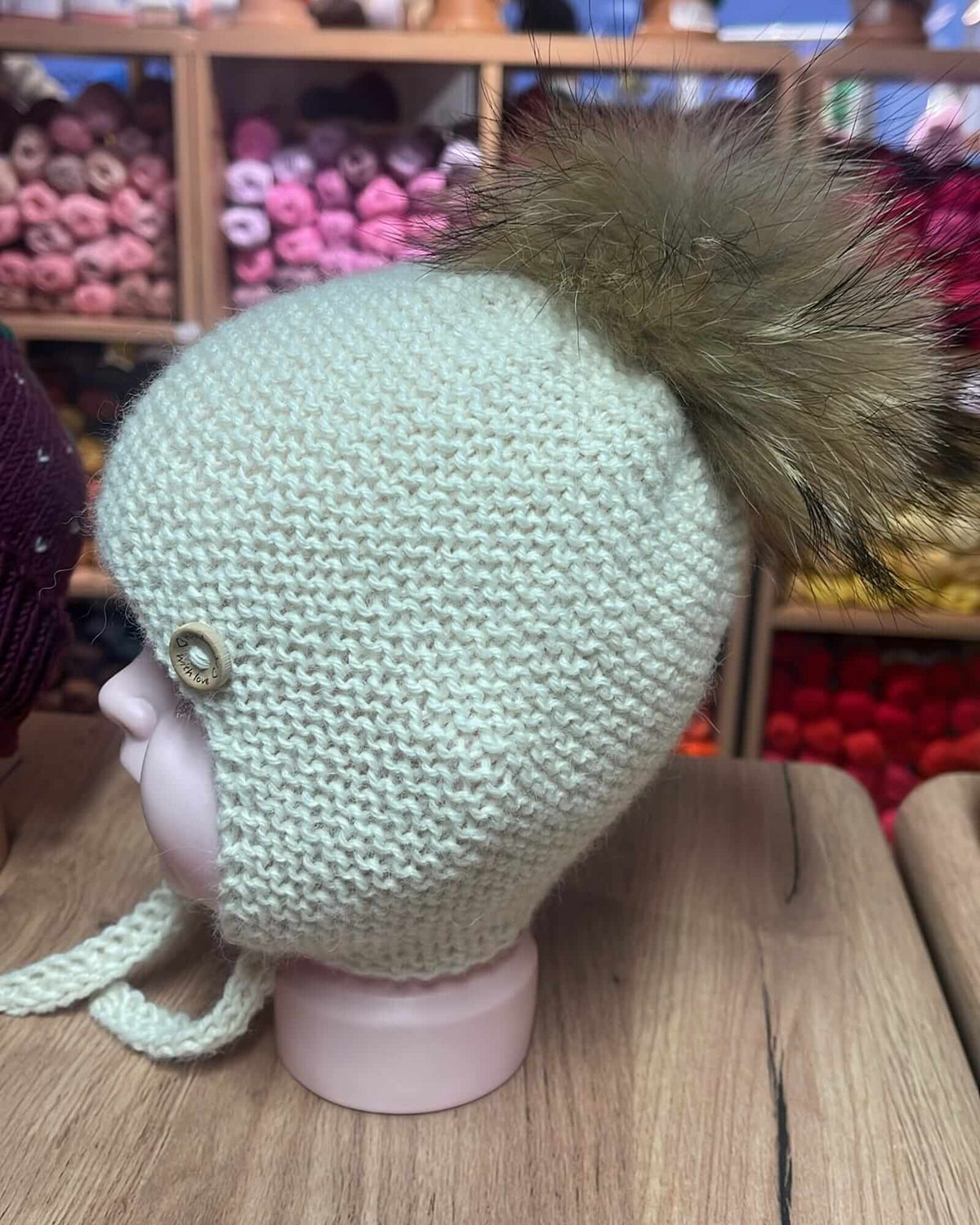 A light sage green knit pom-pom hat with ear flaps and a brown faux fur pom-pom, displayed on a pink mannequin head on a wooden table.