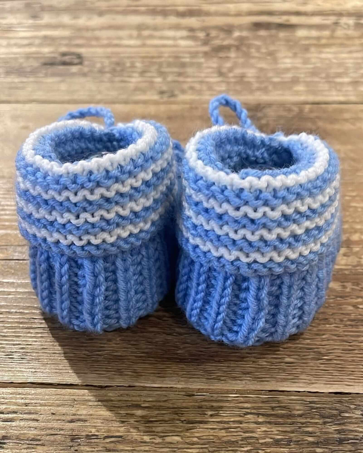 A pair of handmade, hand-knit baby booties in light blue and white stripes, resting on a rustic wooden surface.