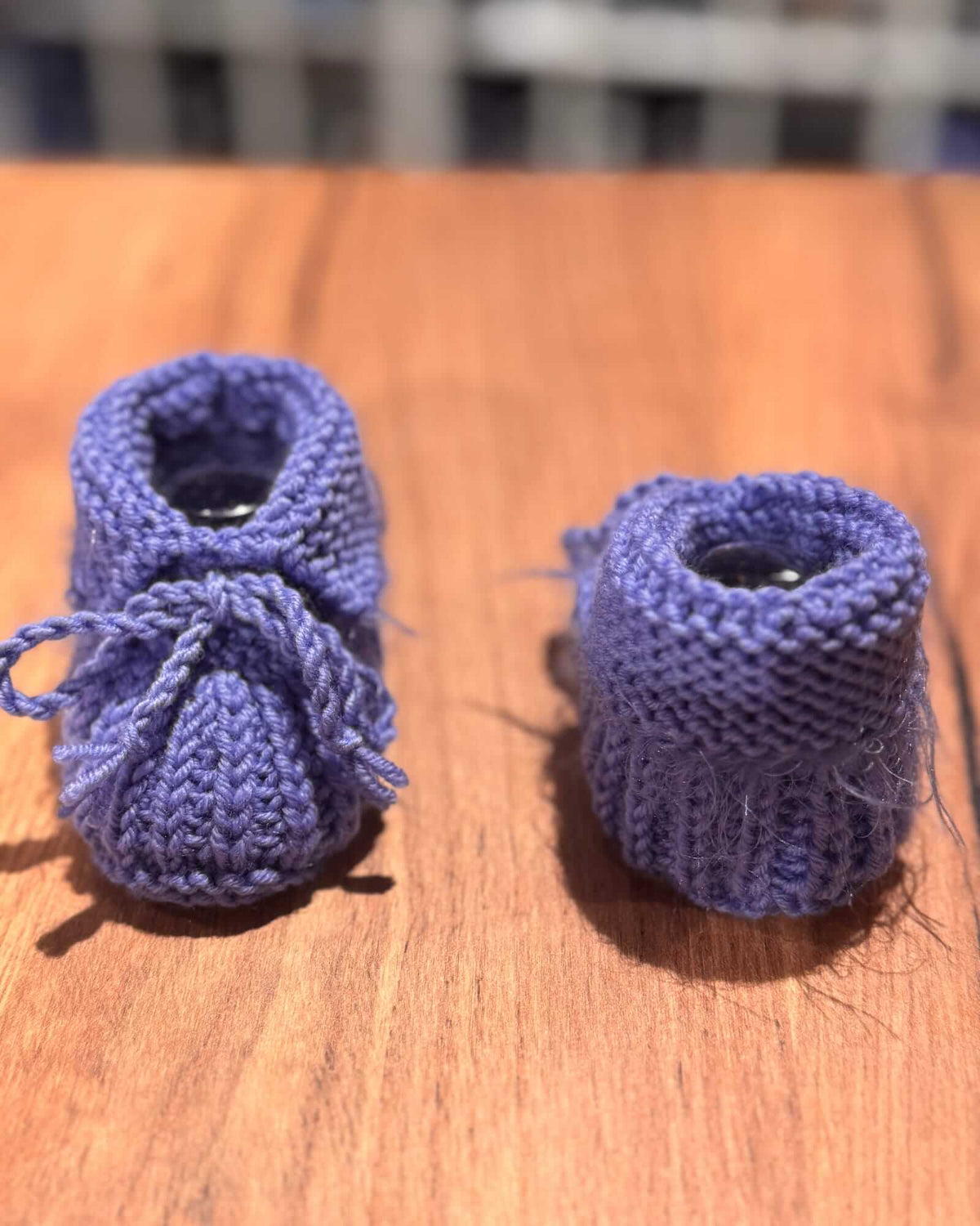 A pair of handmade, hand-knit baby booties in a dusty periwinkle blue, resting on a wooden tabletop with a blurred indoor background.