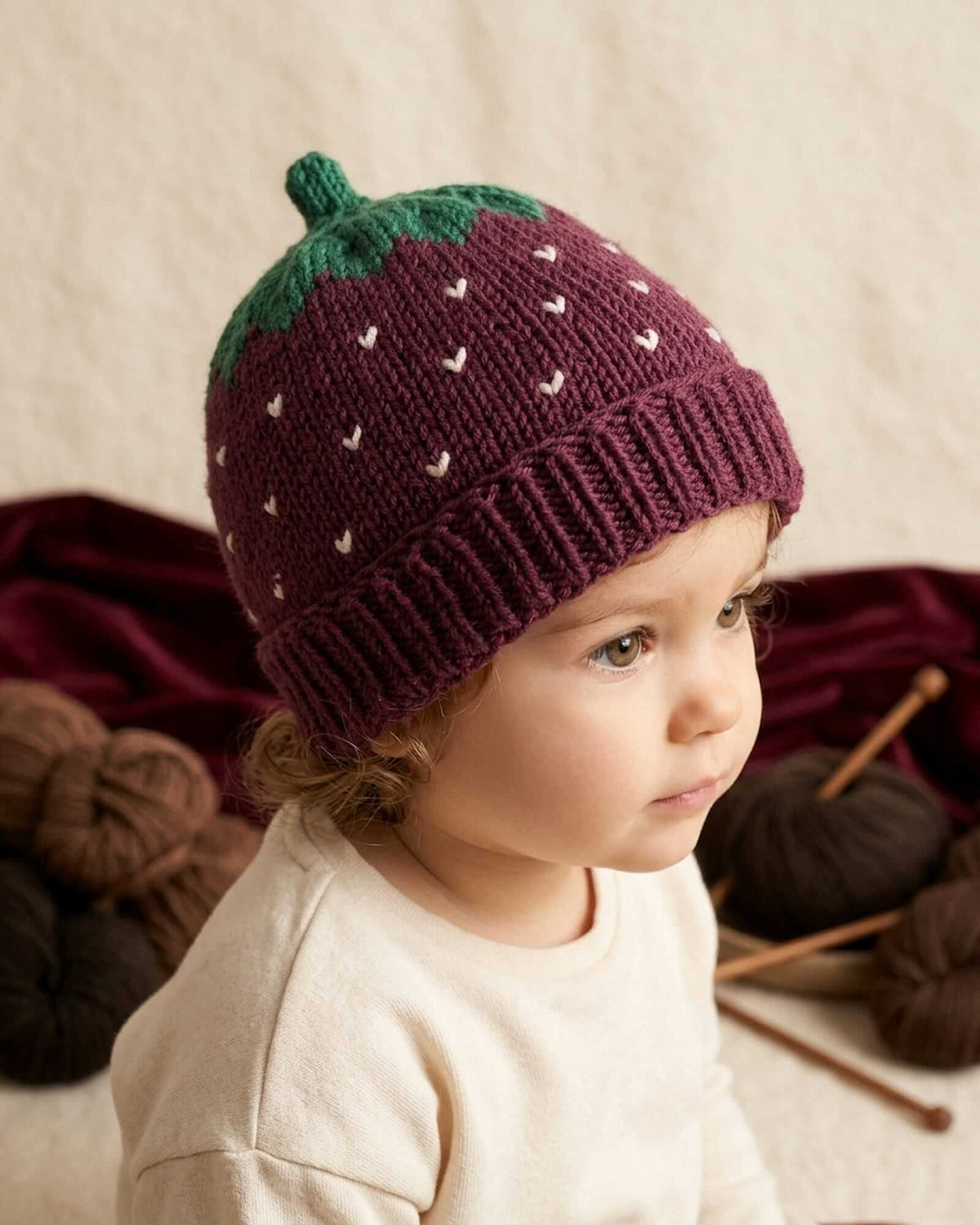 A toddler wearing a knitted strawberry hat in deep purple with a green top and small white leaf details, posed in a cozy setting with yarn balls and knitting needles.