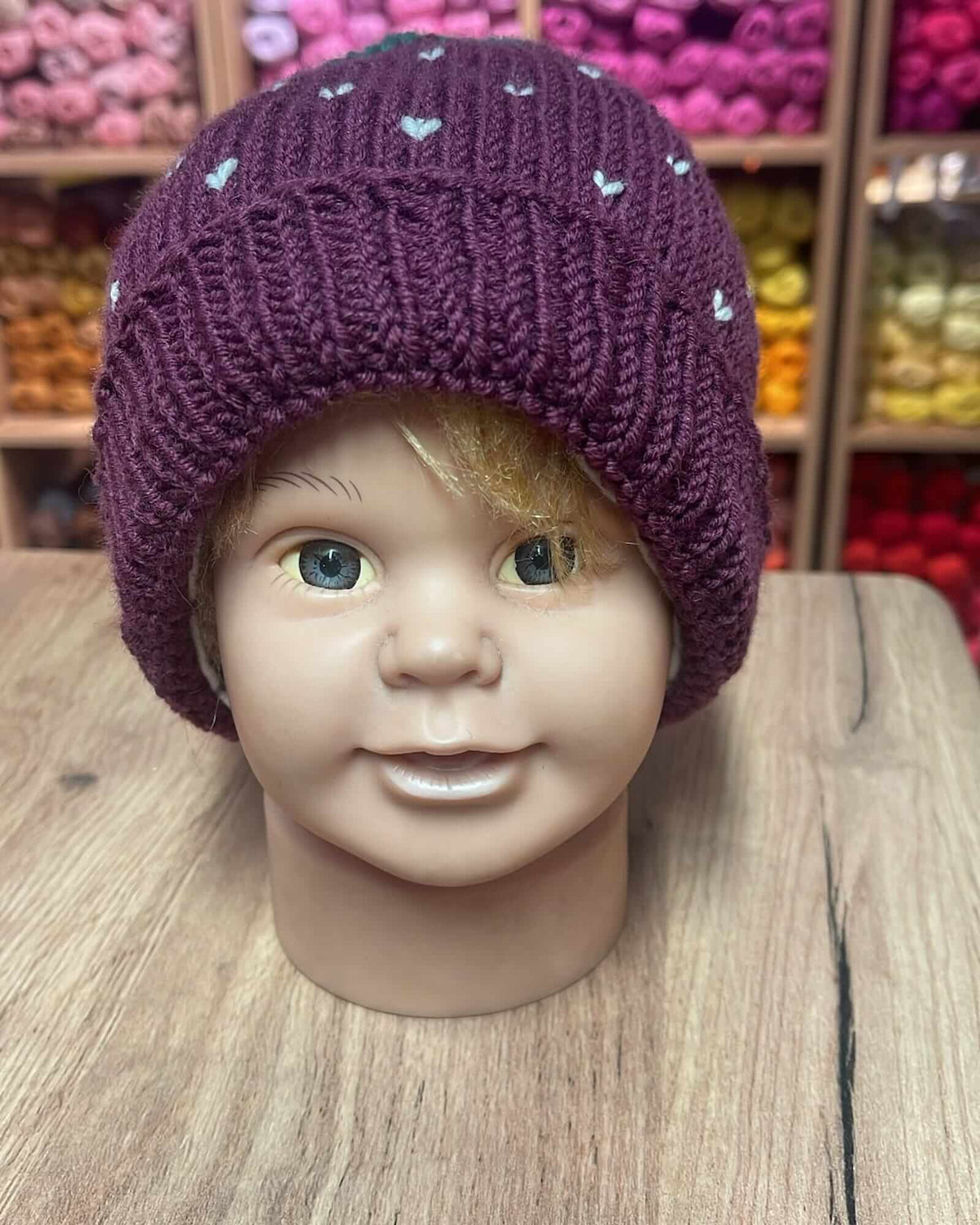 A knitted purple beanie hat with small white heart details, displayed on a mannequin head on a wooden table with shelves of colorful yarn in the background.