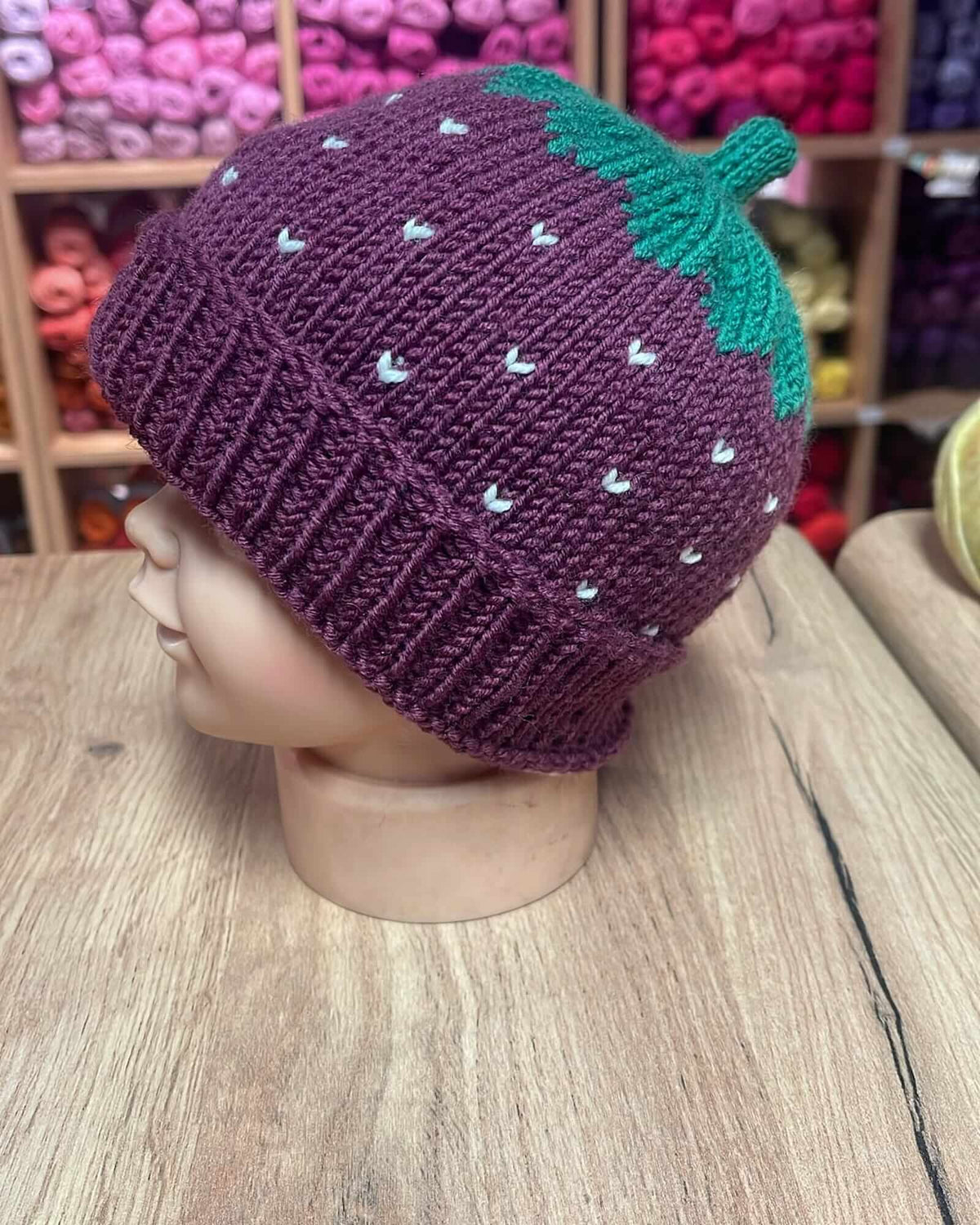 A hand-knit strawberry beanie hat displayed on a mannequin head. The hat is primarily deep purple with a green leafy top and small white embroidered seeds, set against a background of wooden shelves filled with colorful yarn.