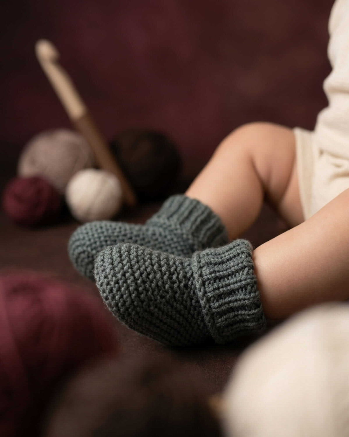 A close-up lifestyle shot of a baby's legs wearing dark gray knit booties, with yarn balls and a crochet hook in the background against a dark maroon backdrop.