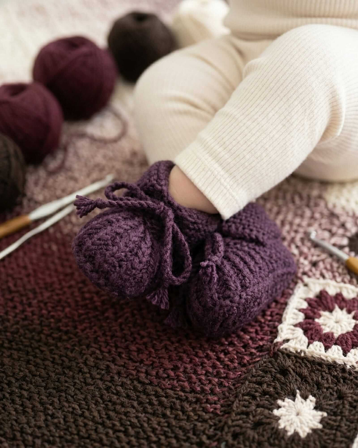 A close-up of a baby's foot wearing a dark purple crocheted bootie, resting on a brown and burgundy crochet blanket with yarn balls and crochet hooks nearby.