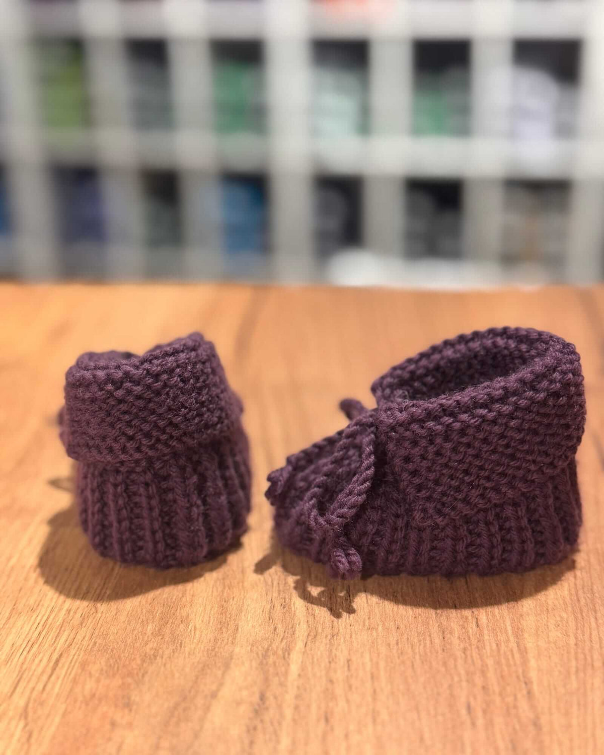 A pair of handmade, hand-knit baby booties in a deep plum purple, displayed on a wooden surface with a blurred background.