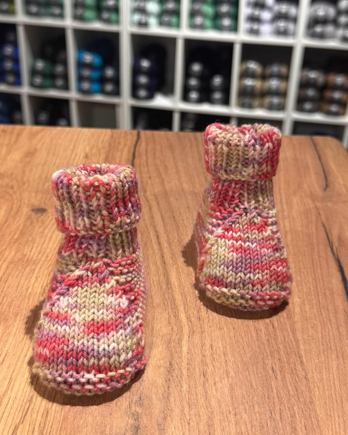 A pair of hand-knit baby booties in a variegated marled yarn of pink, red, beige, and cream, displayed on a wooden tabletop with a blurred background of shelves.