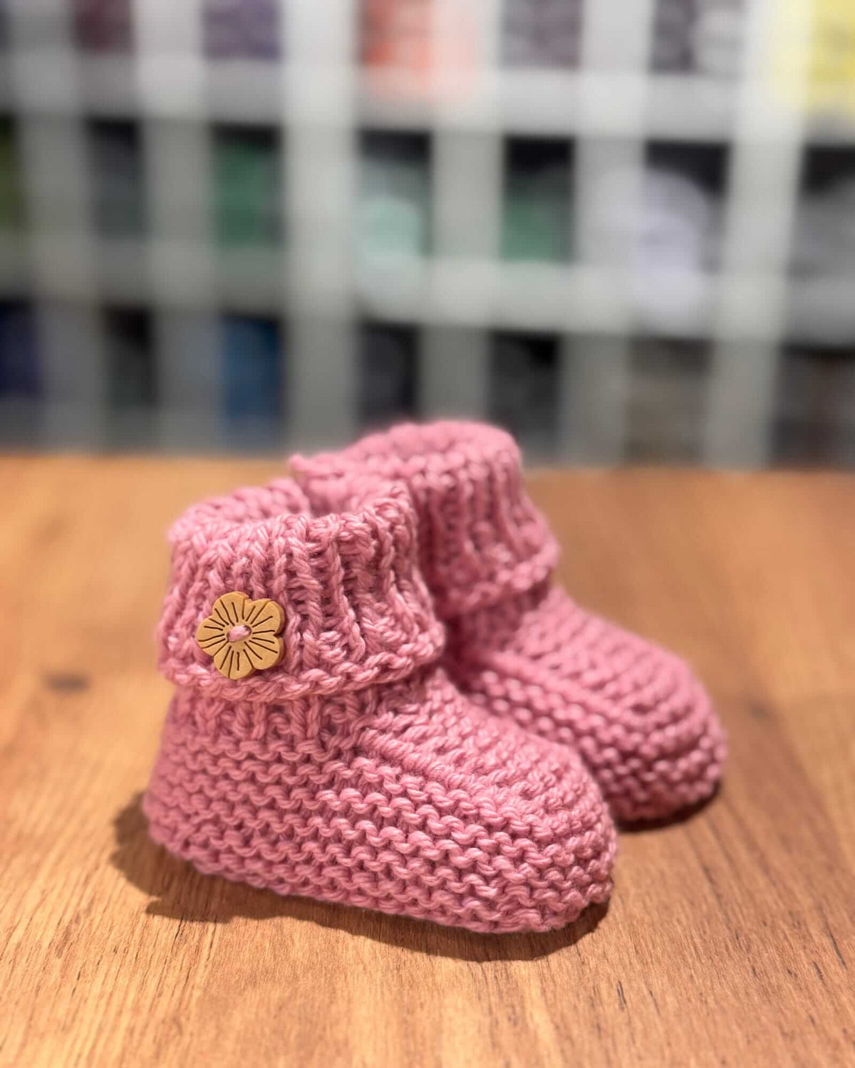 A pair of pink hand-knit baby booties with a small yellow flower accent on the front, resting on a wooden surface with a blurred background.