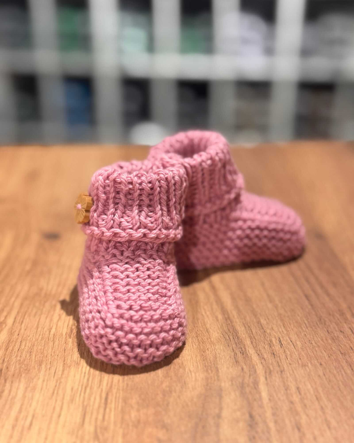 A pair of pink hand-knit baby booties with a small yellow embroidered star detail, resting on a wooden tabletop.