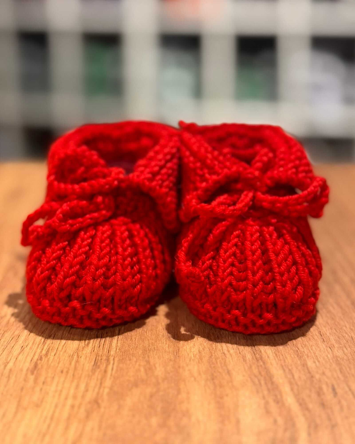 A pair of bright red hand-knitted baby booties with lace-up front details, resting on a wooden surface with a blurred indoor background.