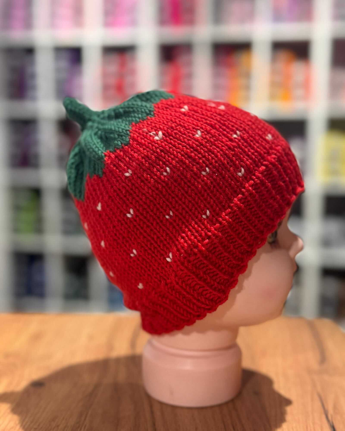 A red knitted strawberry hat with a green leafy top, displayed on a mannequin head stand on a wooden table with a blurred store background.