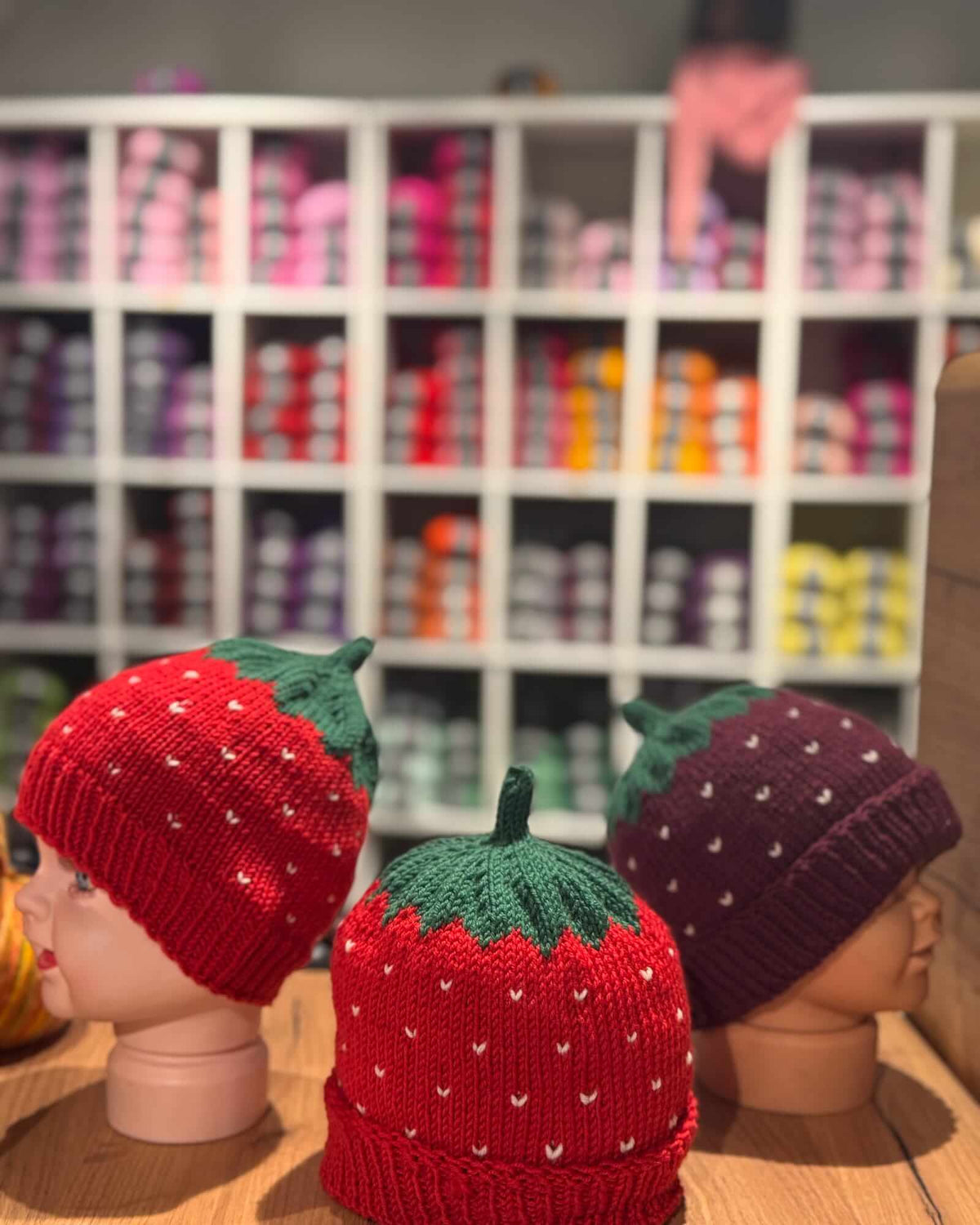 Three hand-knit strawberry-themed baby hats displayed on mannequin heads in a store setting. The center hat is red with green top and white seed details, flanked by a red hat on the left and a dark purple hat on the right, with a blurred shelf of colorful hats in the background.