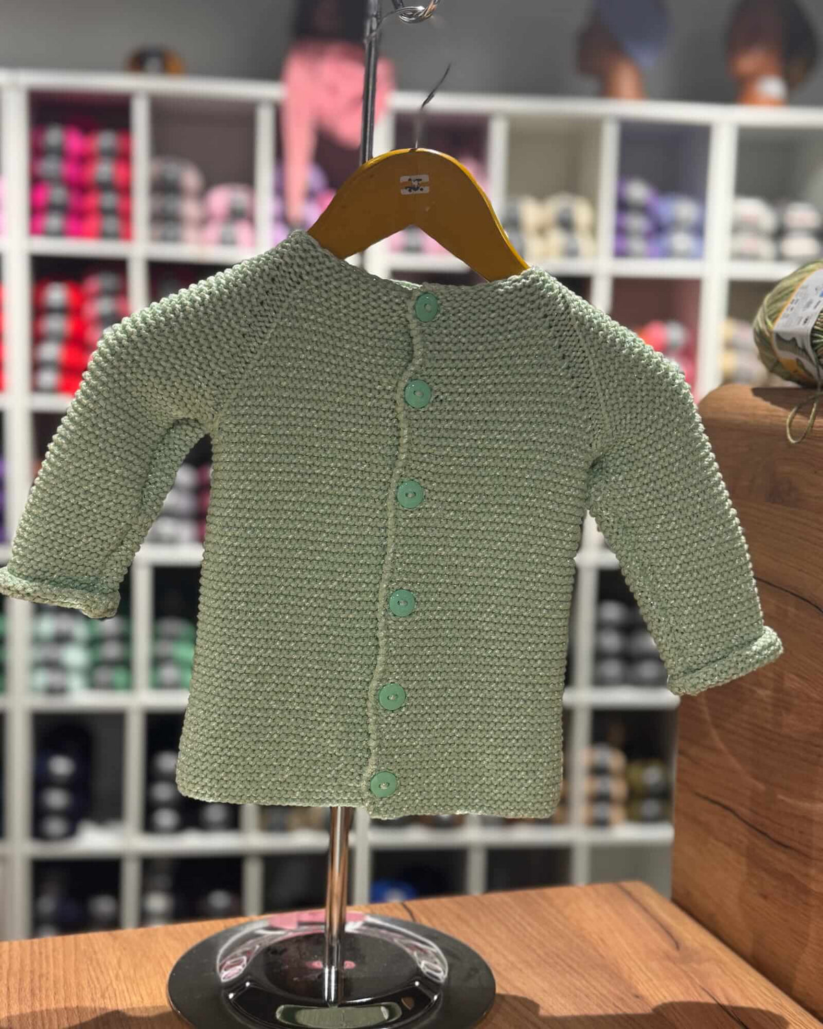 A light sage green knitted baby cardigan with three-quarter sleeves and a button-up front, displayed on a wooden hanger on a stand.