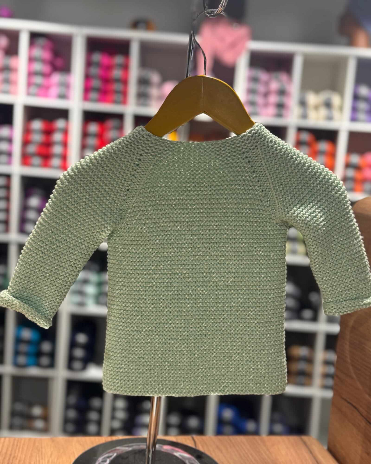 A light sage green knit short-sleeve top displayed on a wooden hanger on a dress form stand, with a blurred store background of shelves containing colorful items.